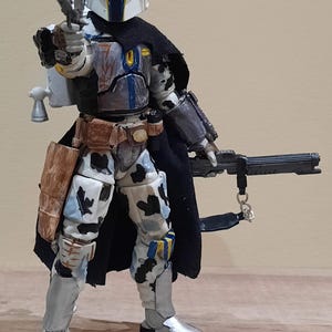 May include: A detailed action figure of a Mandalorian warrior, featuring a white helmet with blue and yellow accents. The figure is clad in armor with a black cape, holding a blaster pistol and a rifle. The armor has a camouflage pattern.