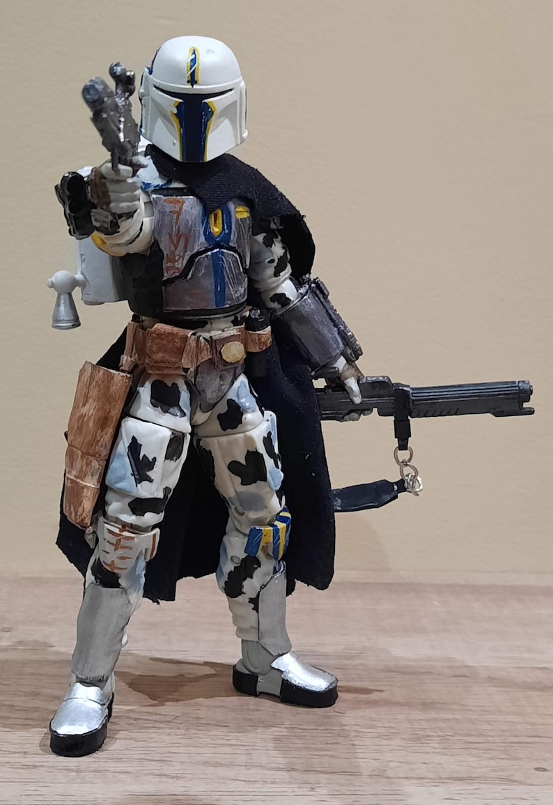 May include: A detailed action figure of a Mandalorian warrior, featuring a white helmet with blue and yellow accents. The figure is clad in armor with a black cape, holding a blaster pistol and a rifle. The armor has a camouflage pattern.