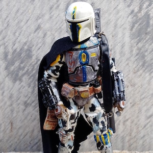May include: A detailed action figure of a Mandalorian warrior in battle armor. The figure features a white helmet with yellow and blue accents, a black cape, and a weathered, multi-colored suit. The figure is approximately 6 inches tall.