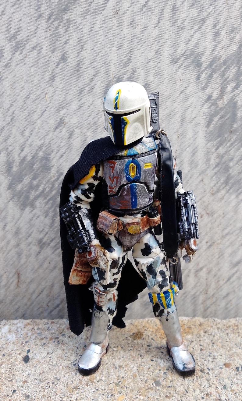 May include: A detailed action figure of a Mandalorian warrior in battle armor. The figure features a white helmet with yellow and blue accents, a black cape, and a weathered, multi-colored suit. The figure is approximately 6 inches tall.