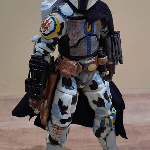 May include: A detailed action figure of a Mandalorian warrior in a white, blue, and black color scheme. The figure has a helmet, cape, and various accessories, including a blaster and a jetpack. The figure is standing on a wooden surface.