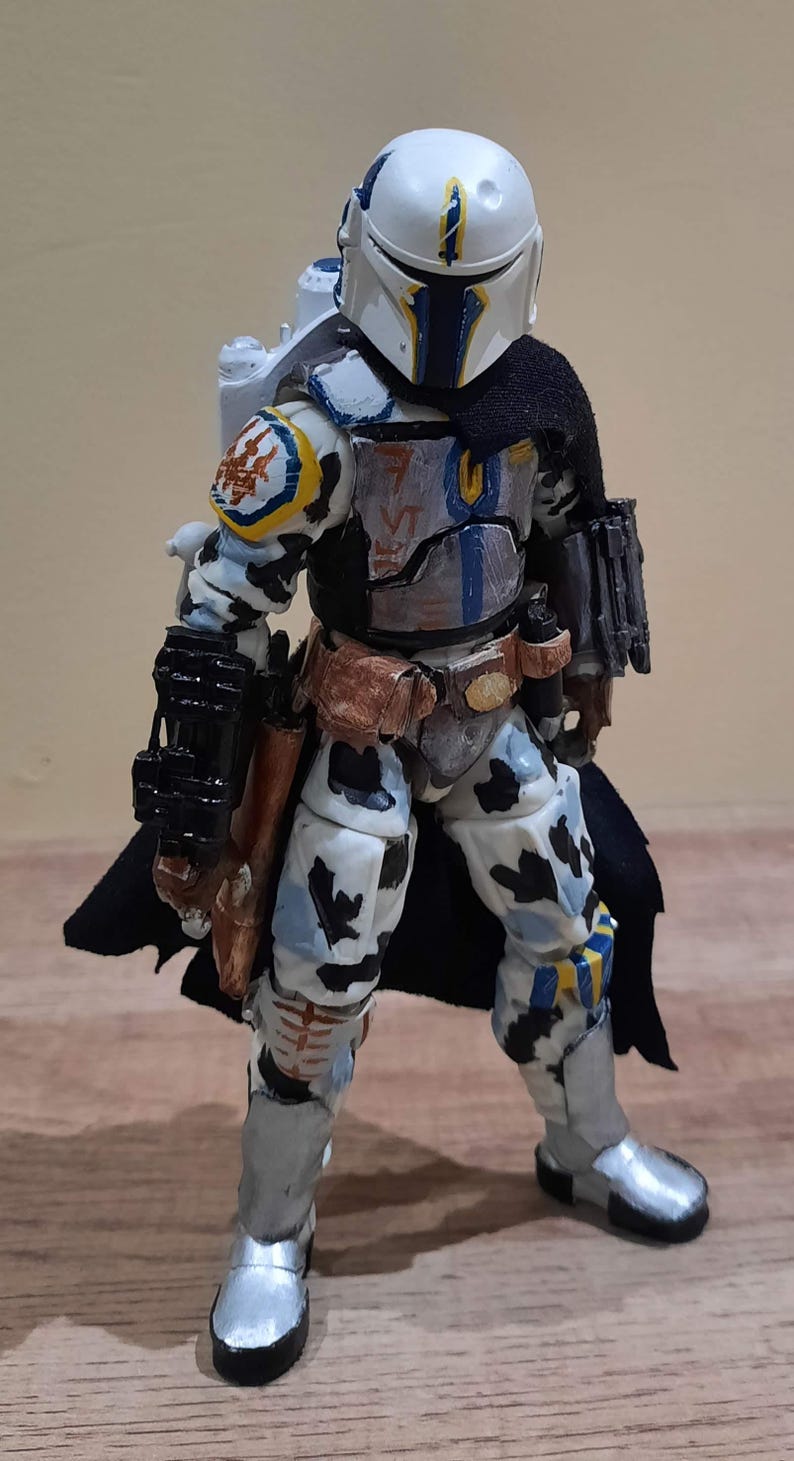 May include: A detailed action figure of a Mandalorian warrior in a white, blue, and black color scheme. The figure has a helmet, cape, and various accessories, including a blaster and a jetpack. The figure is standing on a wooden surface.