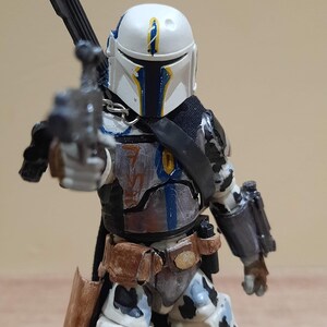 May include: A detailed action figure of a character in a white helmet with blue and yellow accents, holding a blaster. The figure has a black cape and armor with brown and gray details. The figure is approximately 6 inches tall.