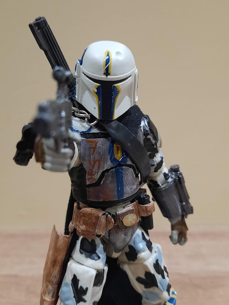 May include: A detailed action figure of a character in a white helmet with blue and yellow accents, holding a blaster. The figure has a black cape and armor with brown and gray details. The figure is approximately 6 inches tall.