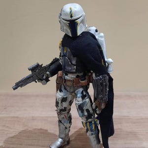 May include: A detailed action figure of a Mandalorian warrior in a battle-worn suit. The figure features a white helmet with blue and yellow accents, a black cape, and a blaster rifle. The armor has a camouflage pattern with silver boots and a jetpack.