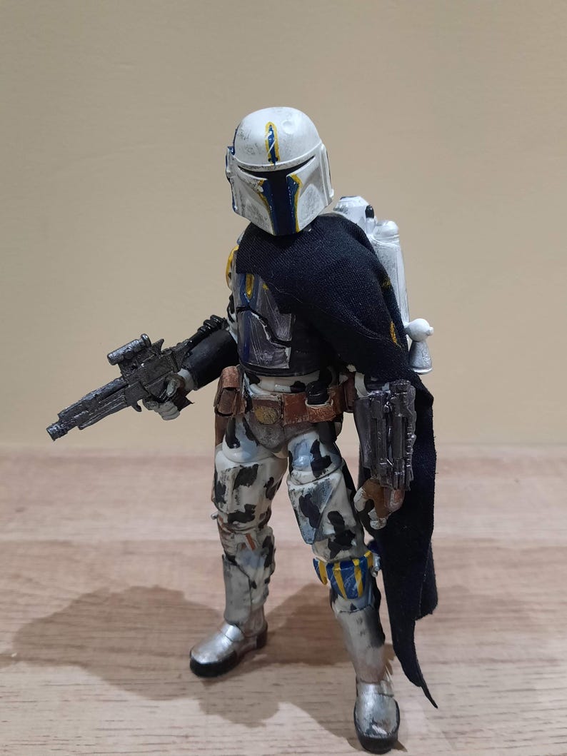 May include: A detailed action figure of a Mandalorian warrior in a battle-worn suit. The figure features a white helmet with blue and yellow accents, a black cape, and a blaster rifle. The armor has a camouflage pattern with silver boots and a jetpack.