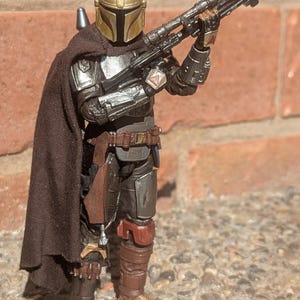 K&ouml;nnte beinhalten: Detaillierte Actionfigur eines mandalorianischen Kriegers, gekleidet in metallischer R&uuml;stung mit goldenem Helm. Die Figur h&auml;lt ein schwarzes Gewehr und tr&auml;gt einen braunen Umhang. Die Figur steht auf einer Steinoberfl&auml;che.