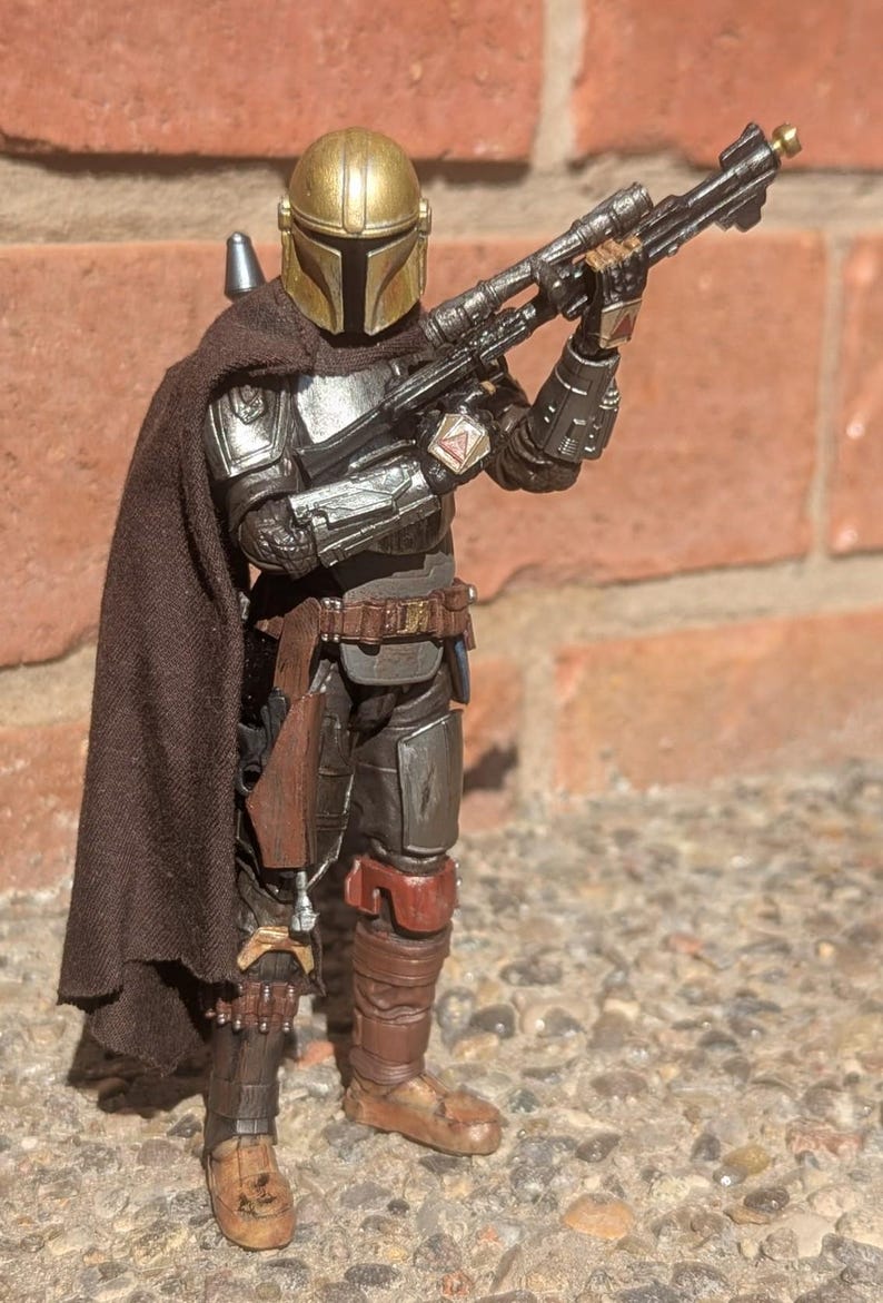 K&ouml;nnte beinhalten: Detaillierte Actionfigur eines mandalorianischen Kriegers, gekleidet in metallischer R&uuml;stung mit goldenem Helm. Die Figur h&auml;lt ein schwarzes Gewehr und tr&auml;gt einen braunen Umhang. Die Figur steht auf einer Steinoberfl&auml;che.