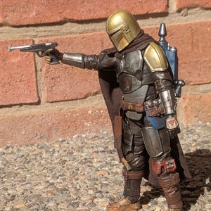 K&ouml;nnte beinhalten: Detaillierte Actionfigur eines Mandalorianer-Charakters, gekleidet in verwitterter R&uuml;stung mit goldenem Helm. Die Figur h&auml;lt eine Blasterpistole, mit braunem Umhang und blauem Jetpack. Die Figur steht auf einer Steinoberfl&auml;che.