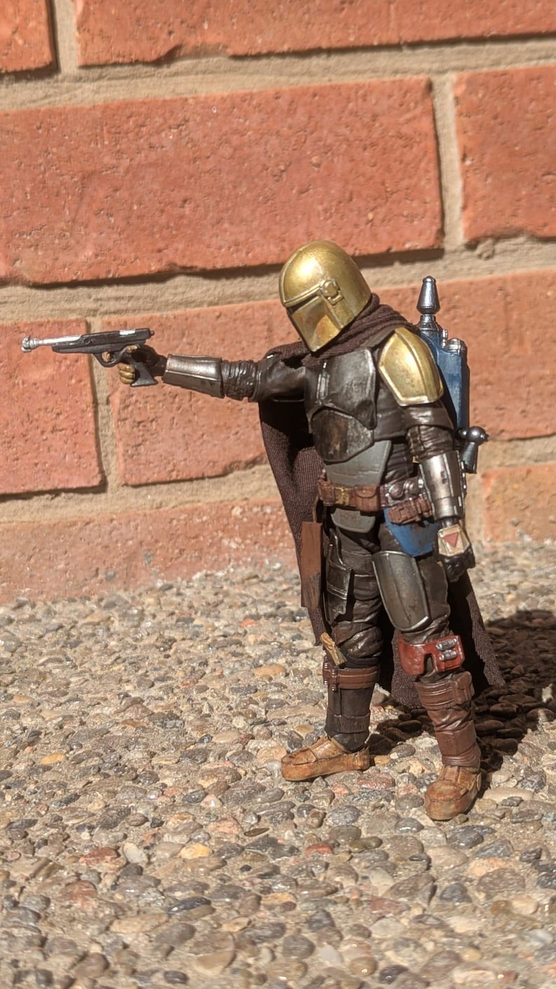 K&ouml;nnte beinhalten: Detaillierte Actionfigur eines Mandalorianer-Charakters, gekleidet in verwitterter R&uuml;stung mit goldenem Helm. Die Figur h&auml;lt eine Blasterpistole, mit braunem Umhang und blauem Jetpack. Die Figur steht auf einer Steinoberfl&auml;che.