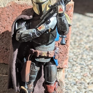 K&ouml;nnte beinhalten: Detaillierte Actionfigur eines mandalorianischen Kriegers, gekleidet in metallischer R&uuml;stung mit goldenem Helm. Die Figur h&auml;lt ein Blastergewehr und posiert vor einer Backsteinmauer. Die Figur hat einen braunen Umhang und tr&auml;gt braune Stiefel.