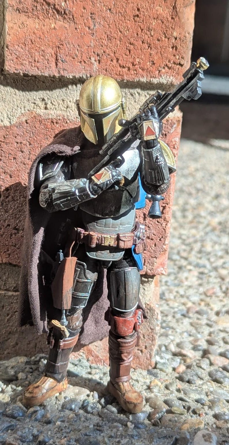K&ouml;nnte beinhalten: Detaillierte Actionfigur eines mandalorianischen Kriegers, gekleidet in metallischer R&uuml;stung mit goldenem Helm. Die Figur h&auml;lt ein Blastergewehr und posiert vor einer Backsteinmauer. Die Figur hat einen braunen Umhang und tr&auml;gt braune Stiefel.