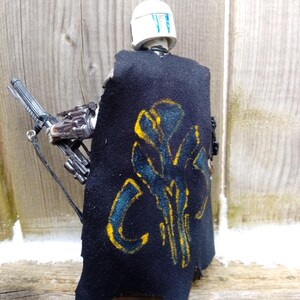 May include: A black cape with a yellow and blue design is worn by a figure with a white helmet. The figure is holding a silver weapon. The cape has a distressed look. The background is a wooden surface.