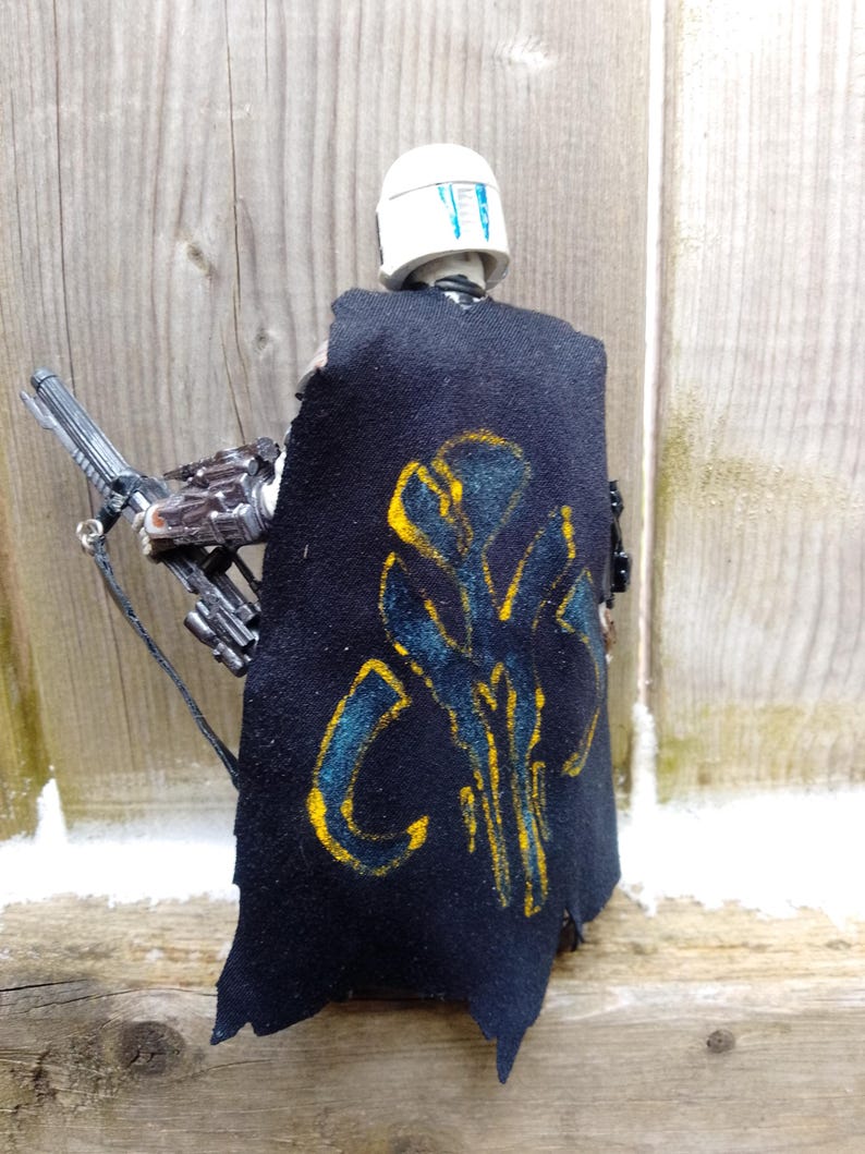 May include: A black cape with a yellow and blue design is worn by a figure with a white helmet. The figure is holding a silver weapon. The cape has a distressed look. The background is a wooden surface.