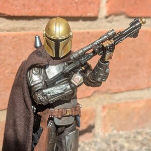 K&ouml;nnte beinhalten: Detaillierte Actionfigur eines mandalorianischen Kriegers, gekleidet in silberner und dunkelbrauner R&uuml;stung, mit einem goldenen Helm. Die Figur h&auml;lt ein schwarzes Gewehr mit Zielfernrohr. Der Hintergrund ist eine rote Backsteinmauer.