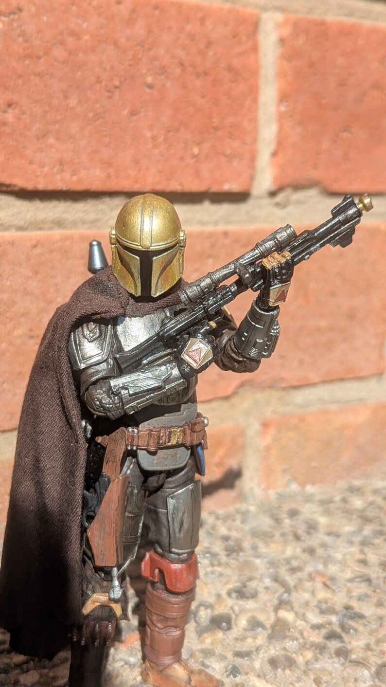 K&ouml;nnte beinhalten: Detaillierte Actionfigur eines mandalorianischen Kriegers, gekleidet in silberner und dunkelbrauner R&uuml;stung, mit einem goldenen Helm. Die Figur h&auml;lt ein schwarzes Gewehr mit Zielfernrohr. Der Hintergrund ist eine rote Backsteinmauer.