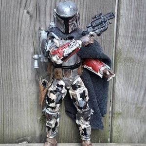 K&ouml;nnte beinhalten: Eine verwitterte, silberne und schwarze Actionfigur eines Kopfgeldj&auml;gers, der einen Helm und eine Tarnung tr&auml;gt. Die Figur h&auml;lt einen Blaster in einer Hand und hat einen Umhang &uuml;ber die Schultern gelegt.