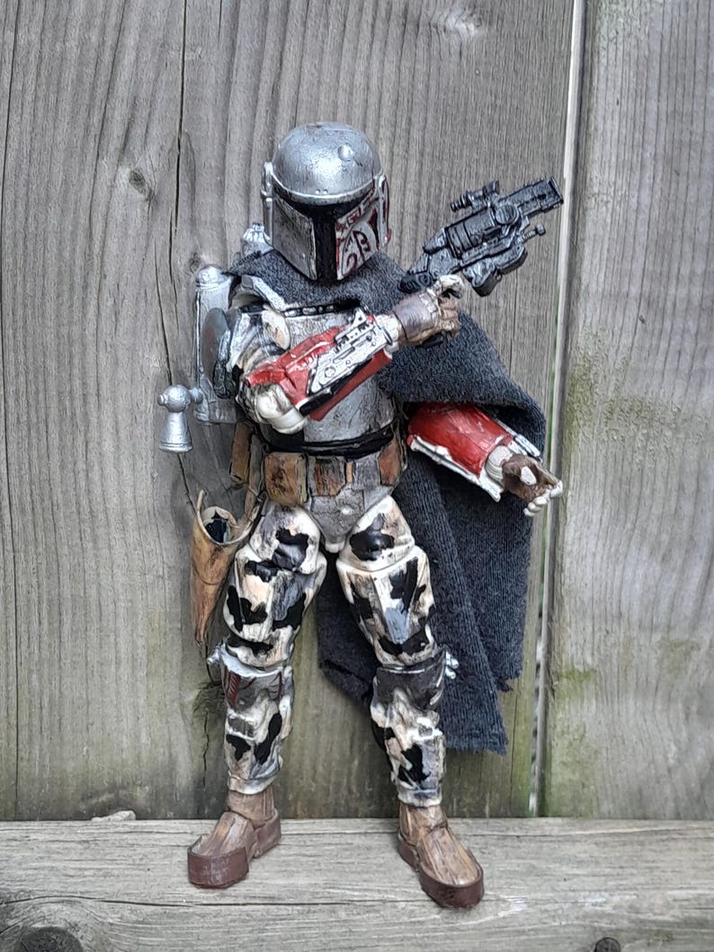 K&ouml;nnte beinhalten: Eine verwitterte, silberne und schwarze Actionfigur eines Kopfgeldj&auml;gers, der einen Helm und eine Tarnung tr&auml;gt. Die Figur h&auml;lt einen Blaster in einer Hand und hat einen Umhang &uuml;ber die Schultern gelegt.
