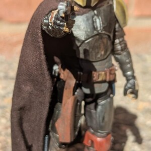 K&ouml;nnte beinhalten: Detaillierte Actionfigur einer Figur aus einem Science-Fiction-Franchise. Die Figur ist in metallischer R&uuml;stung mit einem goldenen Helm und einem braunen Umhang gekleidet. Sie h&auml;lt eine Blasterpistole mit brauner Halfter und Stiefeln.