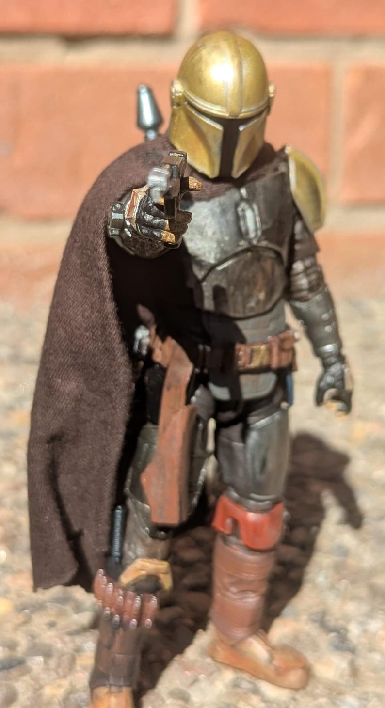 K&ouml;nnte beinhalten: Detaillierte Actionfigur einer Figur aus einem Science-Fiction-Franchise. Die Figur ist in metallischer R&uuml;stung mit einem goldenen Helm und einem braunen Umhang gekleidet. Sie h&auml;lt eine Blasterpistole mit brauner Halfter und Stiefeln.