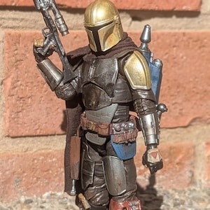 K&ouml;nnte beinhalten: Detaillierte Actionfigur eines Mandalorianischen Kriegers, gekleidet in abgenutzter R&uuml;stung mit goldenem Helm und Schulterpolstern. Die Figur h&auml;lt ein Blastergewehr und verf&uuml;gt &uuml;ber einen Jetpack, was ein Science-Fiction-Thema zeigt.