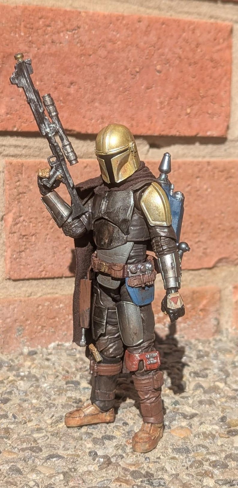 K&ouml;nnte beinhalten: Detaillierte Actionfigur eines Mandalorianischen Kriegers, gekleidet in abgenutzter R&uuml;stung mit goldenem Helm und Schulterpolstern. Die Figur h&auml;lt ein Blastergewehr und verf&uuml;gt &uuml;ber einen Jetpack, was ein Science-Fiction-Thema zeigt.