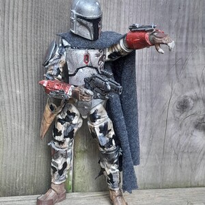 K&ouml;nnte beinhalten: Eine detaillierte Actionfigur eines Mandalorianer-Kriegers, der einen verwitterten, grau-silbernen Helm und eine R&uuml;stung tr&auml;gt. Die Figur h&auml;lt ein Blastergewehr und hat eine rot-schwarze am Handgelenk montierte Waffe. Die Figur steht auf einer Holzoberfl&auml;che.
