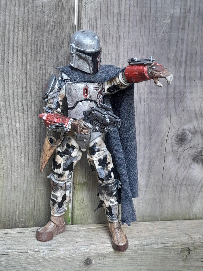 K&ouml;nnte beinhalten: Eine detaillierte Actionfigur eines Mandalorianer-Kriegers, der einen verwitterten, grau-silbernen Helm und eine R&uuml;stung tr&auml;gt. Die Figur h&auml;lt ein Blastergewehr und hat eine rot-schwarze am Handgelenk montierte Waffe. Die Figur steht auf einer Holzoberfl&auml;che.