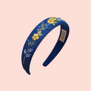 May include: A navy blue headband with embroidered yellow and blue flowers and green leaves. The floral design is on the top of the headband. The headband has a curved shape and a label that says "Ola Mia".
