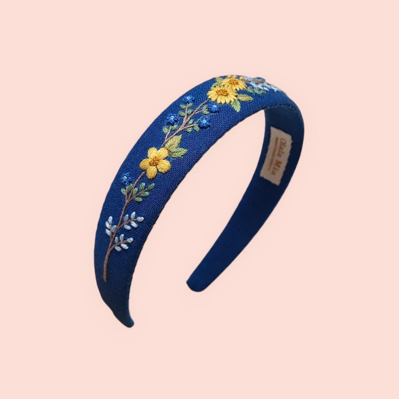 May include: A navy blue headband with embroidered yellow and blue flowers and green leaves. The floral design is on the top of the headband. The headband has a curved shape and a label that says "Ola Mia".