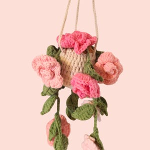 Puede incluir: Un macetero colgante de ganchillo hecho a mano con flores rosas y rosa claro con hojas verdes. El macetero está suspendido por una cuerda beige. Las flores están dispuestas alrededor del macetero, creando un diseño floral decorativo.