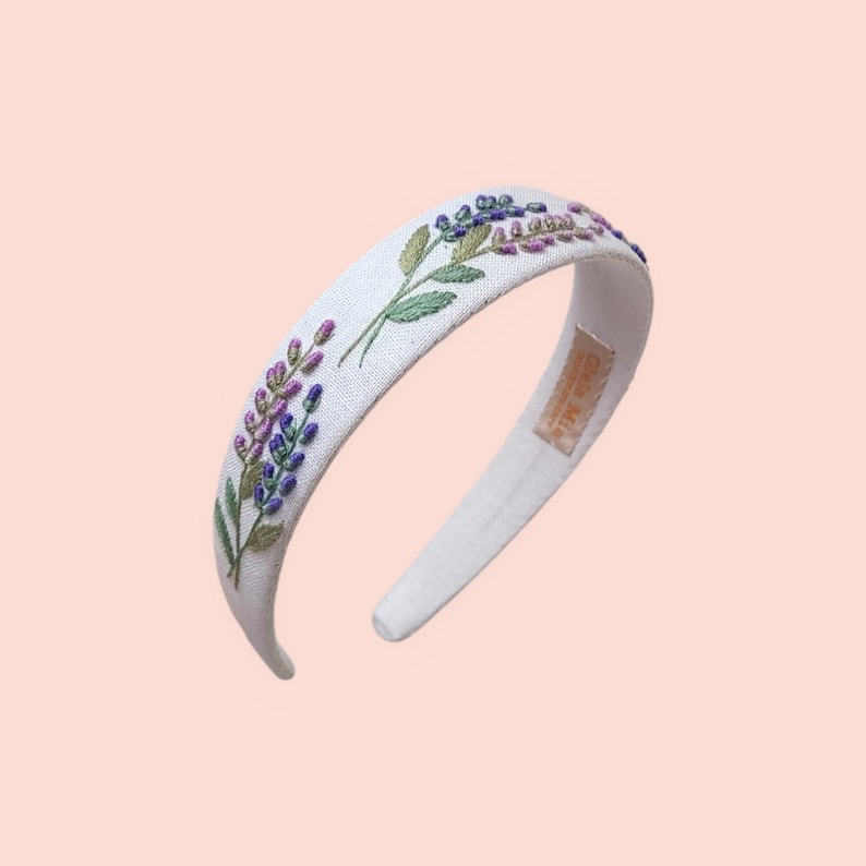 May include: White headband with embroidered lavender sprigs in purple and green. The floral design is on the top of the headband. The headband is made of a soft, textured fabric.