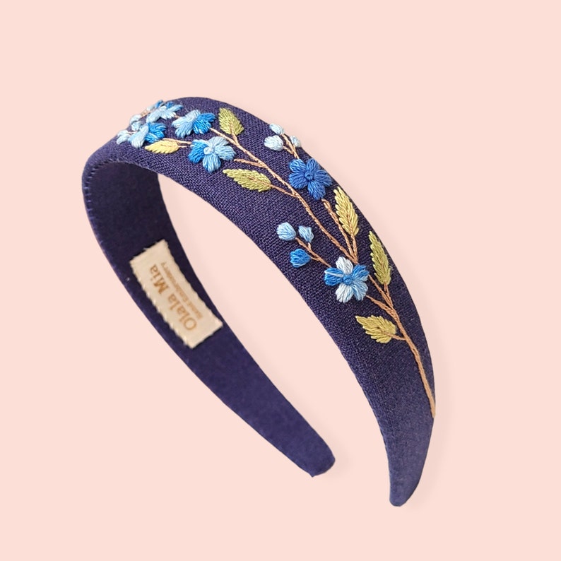 May include: A navy blue headband with embroidered blue flowers and green leaves. The headband has a fabric-covered design and features the text "Ola la Mia" on a small tag.