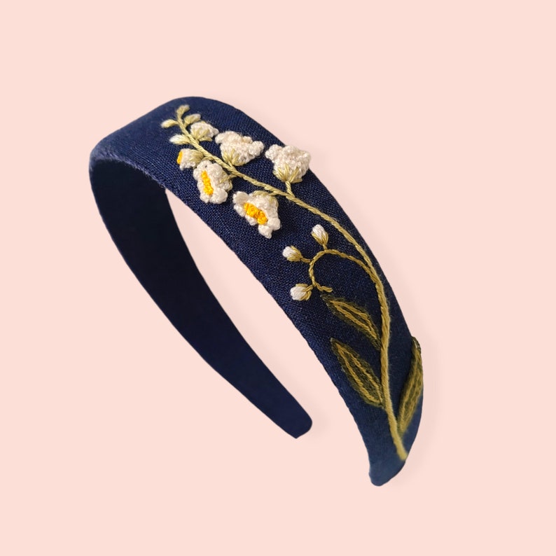 May include: A navy blue headband with embroidered white and yellow flowers and green leaves. The floral design is on the left side of the headband. The headband is made of a soft fabric and is suitable for everyday wear.