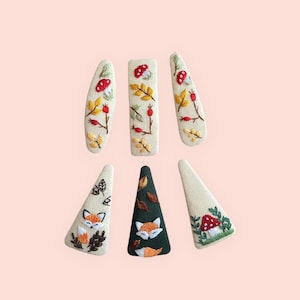 Könnte beinhalten: Sechs bestickte Haarspangen mit Herbstmotiven. Die Spangen zeigen Pilze, Blätter und Füchse in Creme, Grün, Orange und Rot. Sie sind auf einem hellrosa Hintergrund angeordnet. Die Designs sind aufwendig gestickt.