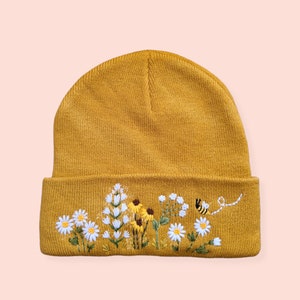 Puede incluir: Gorro mostaza con diseño floral y abeja bordados. Las flores bordadas incluyen margaritas blancas, girasoles amarillos y otras flores silvestres. Una pequeña abeja vuela cerca de las flores. El gorro es de estilo con dobladillo.