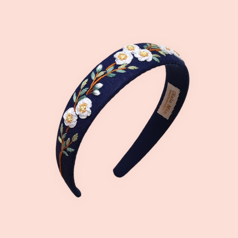 May include: A navy blue headband with embroidered white flowers and green leaves. The floral design is accented with gold thread. The headband is a stylish hair accessory.