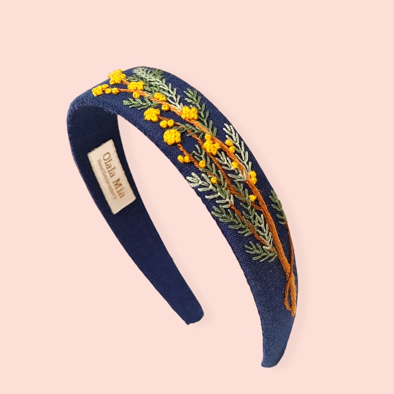 May include: A navy blue headband with embroidered yellow flowers and green leaves. The headband features the text "Olala Mia Hand Embroidery" on a small tag. The floral design is detailed and adds a touch of nature-inspired elegance.