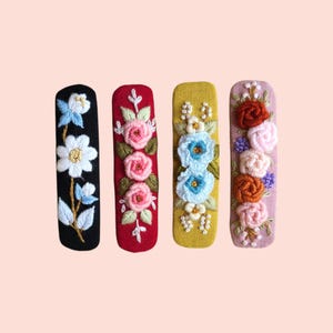 Puede incluir: Cuatro horquillas bordadas en fila. Cada horquilla presenta un color y un diseño floral diferentes. Los colores incluyen negro, rojo, amarillo y rosa. Los diseños florales incluyen rosas, margaritas y otras flores.