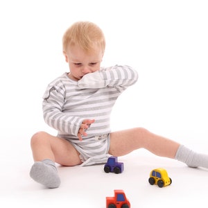 Puede incluir: Un ni&ntilde;o peque&ntilde;o con un body a rayas grises y blancas y calcetines grises est&aacute; sentado sobre una superficie blanca, rodeado de coches de juguete de colores. Los coches son rojos, amarillos, verdes y morados.