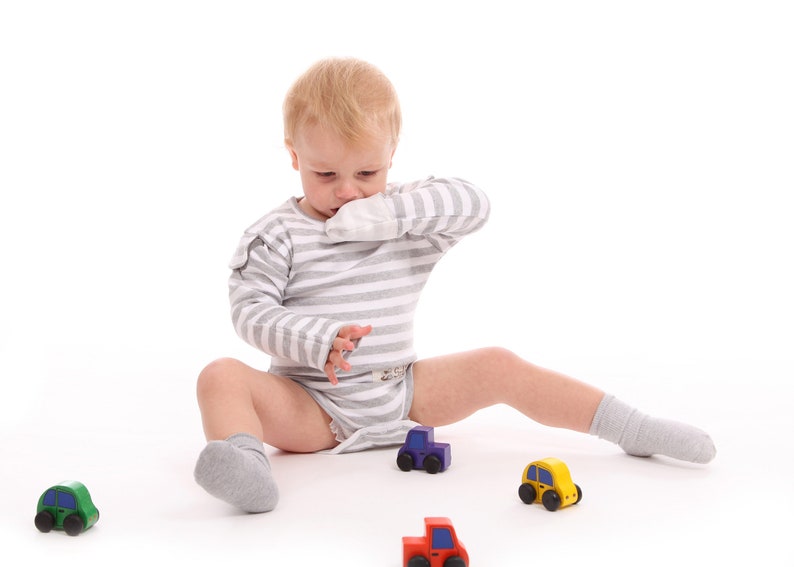 Puede incluir: Un ni&ntilde;o peque&ntilde;o con un body a rayas grises y blancas y calcetines grises est&aacute; sentado sobre una superficie blanca, rodeado de coches de juguete de colores. Los coches son rojos, amarillos, verdes y morados.