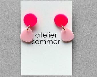 Könnte beinhalten: Ohrringe mit einem leuchtend pinkfarbenen Kreis und einem hellrosa, unregelmäßig geformten Tropfen. Die Ohrringe sind durch einen goldfarbenen Ring verbunden. Der Text "atelier sommer" ist auf eine weiße Karte gedruckt.