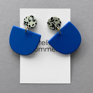 Könnte beinhalten: Ein Paar blaue Statement-Ohrringe mit halbkreisförmiger Form. Die Ohrringe haben eine gesprenkelte, runde Oberseite in Schwarz und Weiß, verbunden durch einen goldfarbenen Ring. Sie werden auf einer weißen Karte präsentiert.