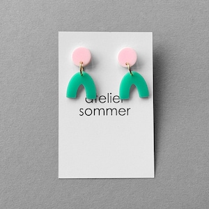 Könnte beinhalten: Ein Paar Ohrringe mit einem rosa Kreis und einer teal-farbenen Bogenform, verbunden durch goldfarbene Haken. Die Ohrringe werden auf einer weißen Karte mit dem Text "atelier sommer" präsentiert.