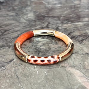 Könnte beinhalten: Ein farbenfrohes Armband mit goldfarbenen Metallakzenten. Das Armband hat Segmente in Orange, Pfirsich und einem gemusterten braun-weißen Design. Das Armband wird auf einer grauen Marmoroberfläche präsentiert.