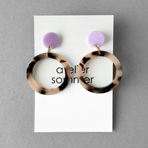 Könnte beinhalten: Ohrringe mit einem hellvioletten Stecker und einem kreisförmigen Reifen mit einem braunen und schwarzen Fleckenmuster. Die Ohrringe hängen an einer weißen Karte mit dem Text "atelier sonner".