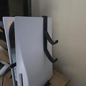 Peut inclure: Une console PlayStation 5 blanche montée sur un mur avec un support vertical noir. La console est sur une surface en bois.