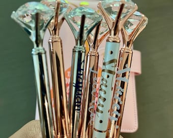 May include: A set of six rose gold pens with clear diamond-shaped tops. Each pen has a different inscription on the barrel, including "Fiercely", "Beautifully", "Wonderfully", "Perfectly", "Beautifully", and "Wonderfully".