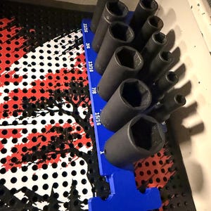 May include: A set of black metal impact sockets arranged on a blue plastic organizer tray. The tray features printed size labels including 11/16, 3/4, 13/16, 7/8, 15/16, and 1 inch. The sockets are cylindrical with hexagonal openings at the top.