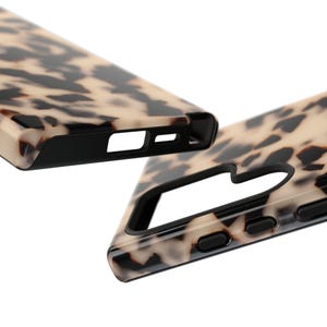 May include: Two smartphone cases featuring a glossy tortoiseshell pattern with a mix of cream, tan, and dark brown spots. The cases have a solid black interior frame with precise cutouts for the camera lens, charging port, and side buttons.